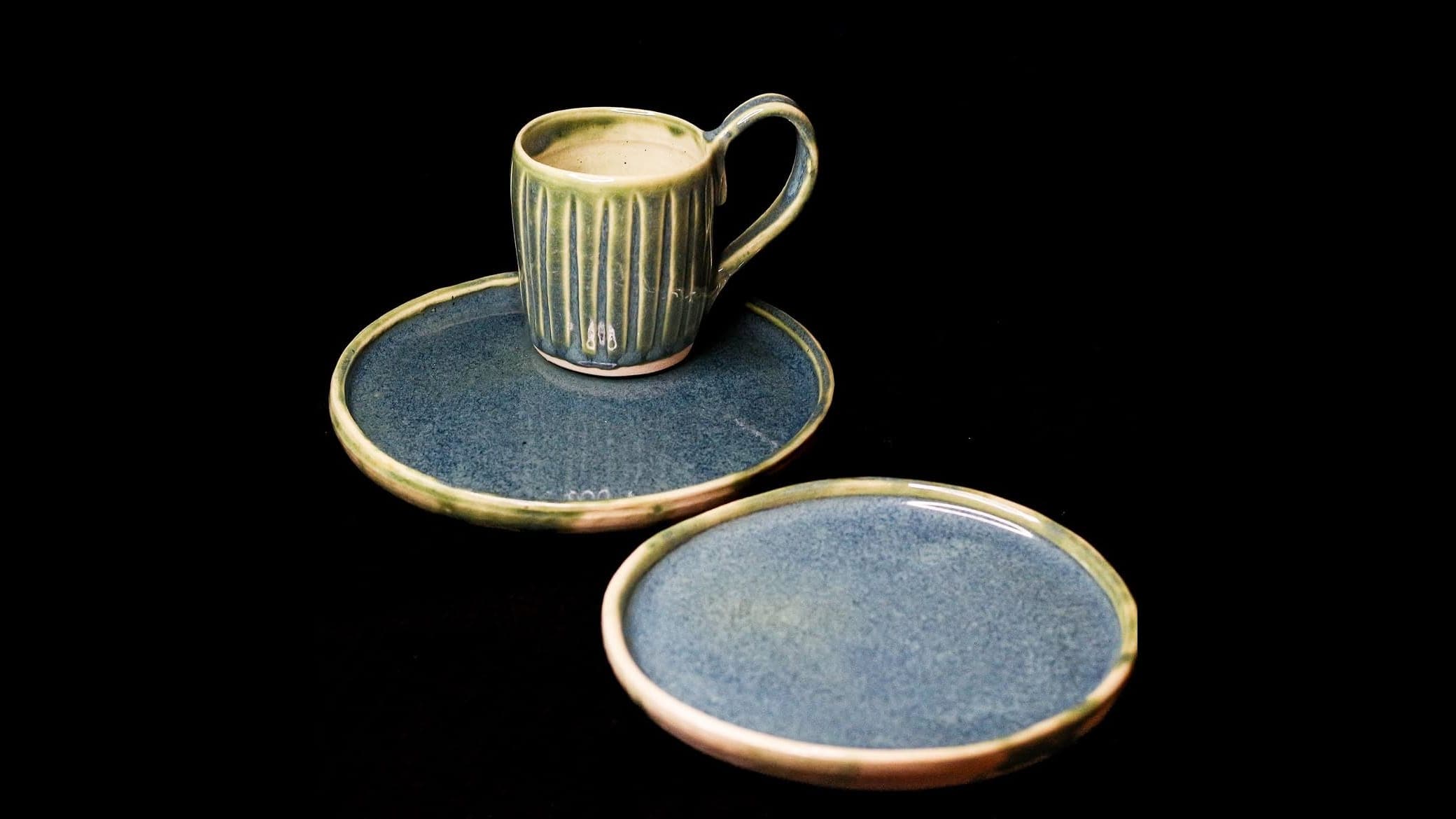 A fluted green-glazed cup on a blue-glazed plate with a second plate beside it, photographed against black.